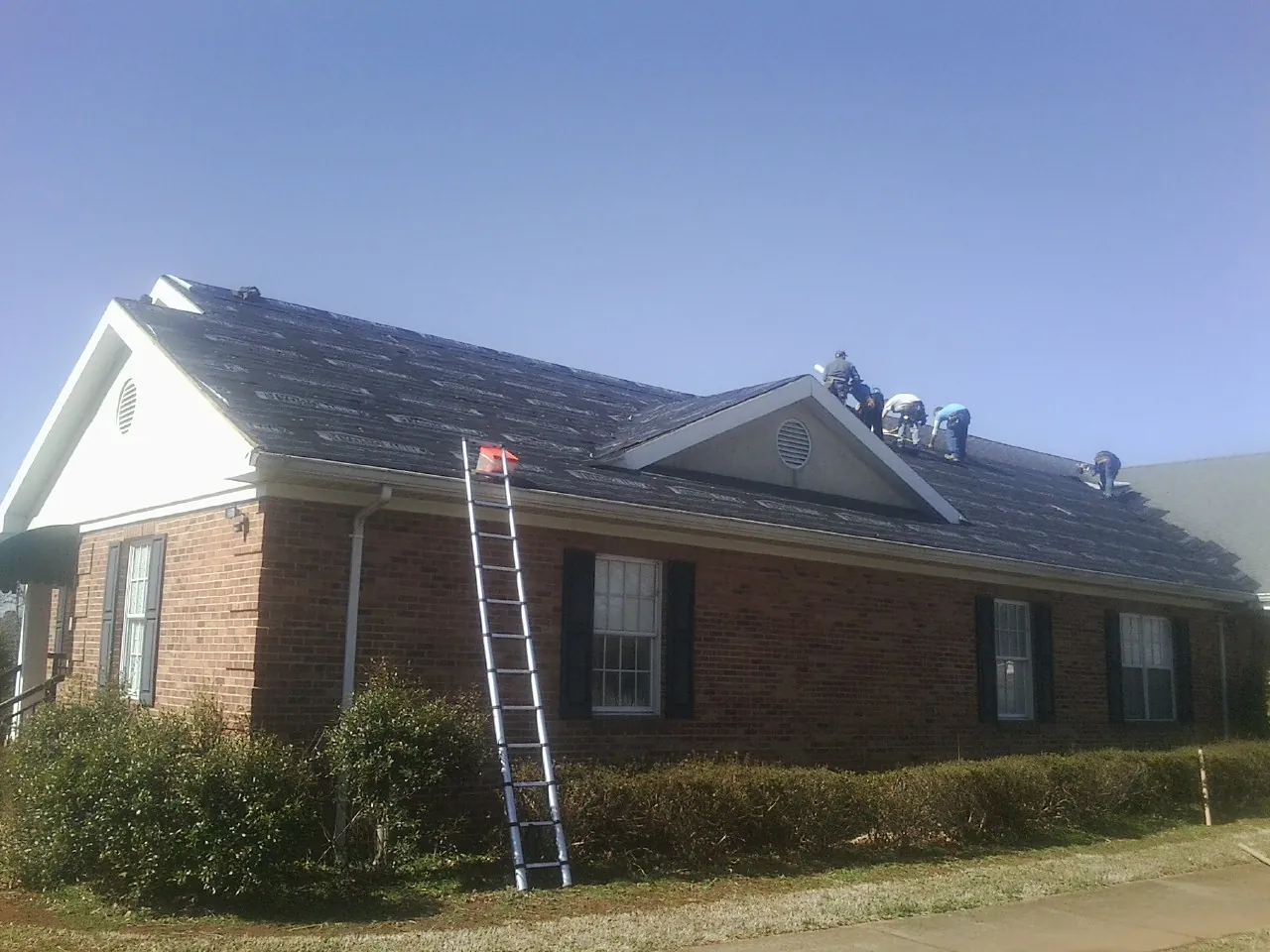 Crew tearing shingles off a brick ranch roof with a ladder set — Metal Roof Repair work by our crew in Gill